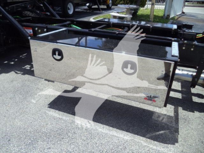 Vehicle chassis with a flatbed toolbox mounted behind the cab; polished metal toolbox with two circular cutouts and a badge on the lower right corner, attached to a truck frame.