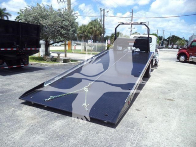 Tow truck rollback flatbed showing a black steel ramp bed with a raised headache rack and rear wheel chocks; blue sky and palm trees in background.