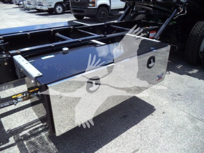 Truck chassis with a shiny rectangular tool/storage box mounted on the frame, hydraulic lines and truck cab visible in background