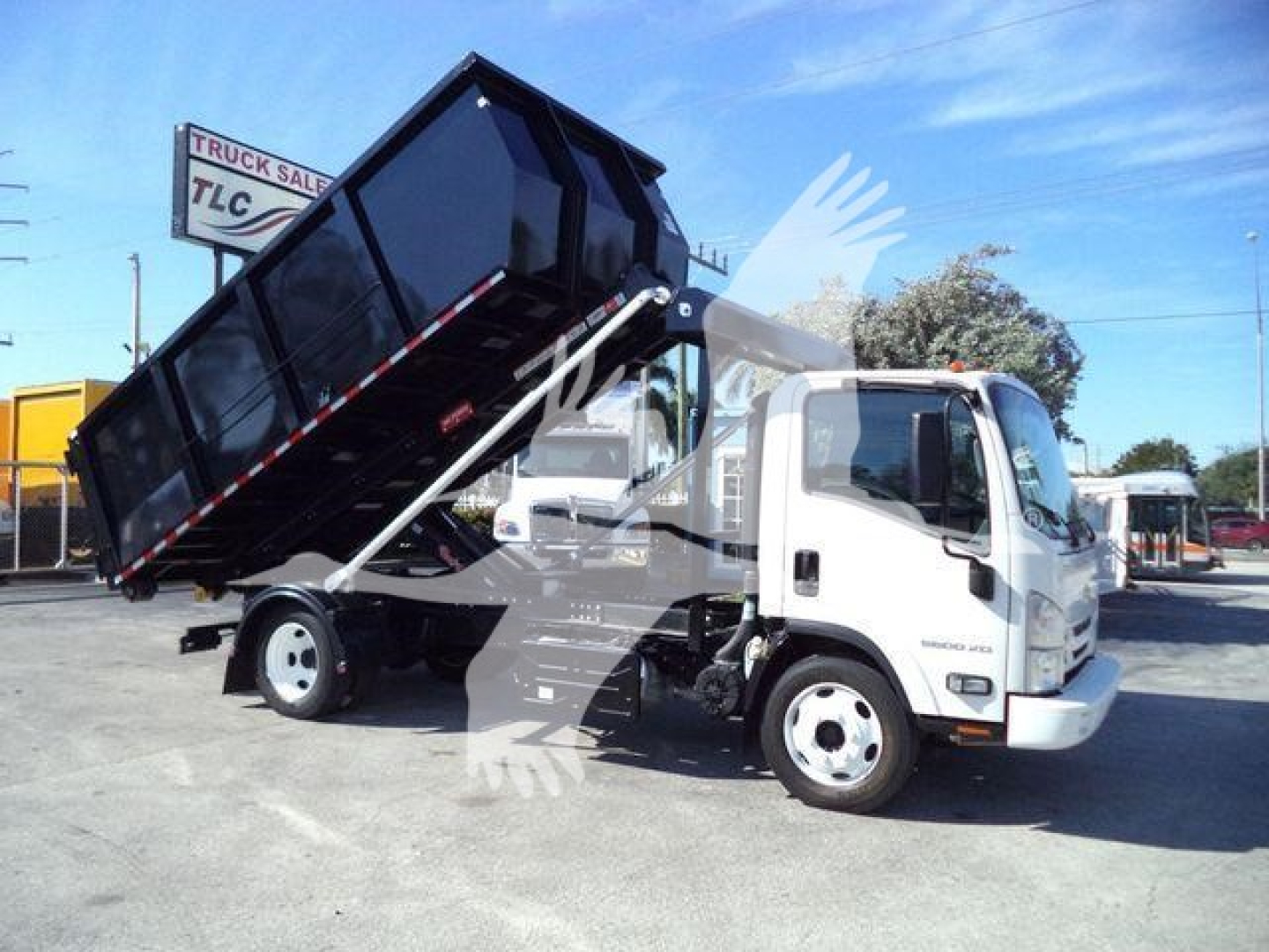 Isuzu NPR/NQR style dump truck with a raised black dump bed on a white cab, visible text includes dealership sign 'TRUCK SALES TLC' in background and No other legible markings.