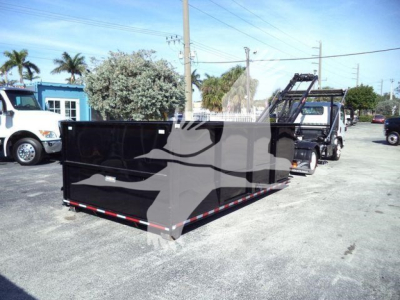Isuzu NPRR truck with a black roll-off container attached, showing a large black cargo bed/dump container and a truck behind in a lot. Visible text: none legible except general scene.