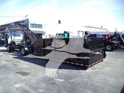 Isuzu NPR/NRR roll-off truck with a large black Dump/box container. In the background are other trucks and a building. No visible labels readable.