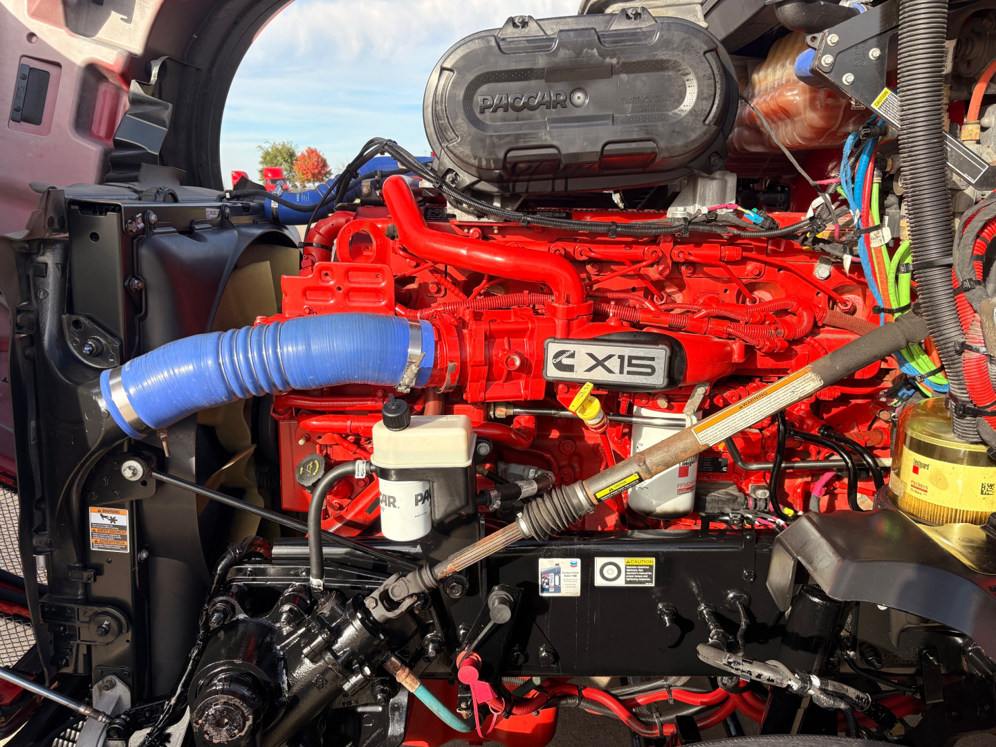 Kenworth T680 engine bay with bright red Cummins/CX15 engine, blue intake hose, white reservoir, and various hoses and electricals visible.
