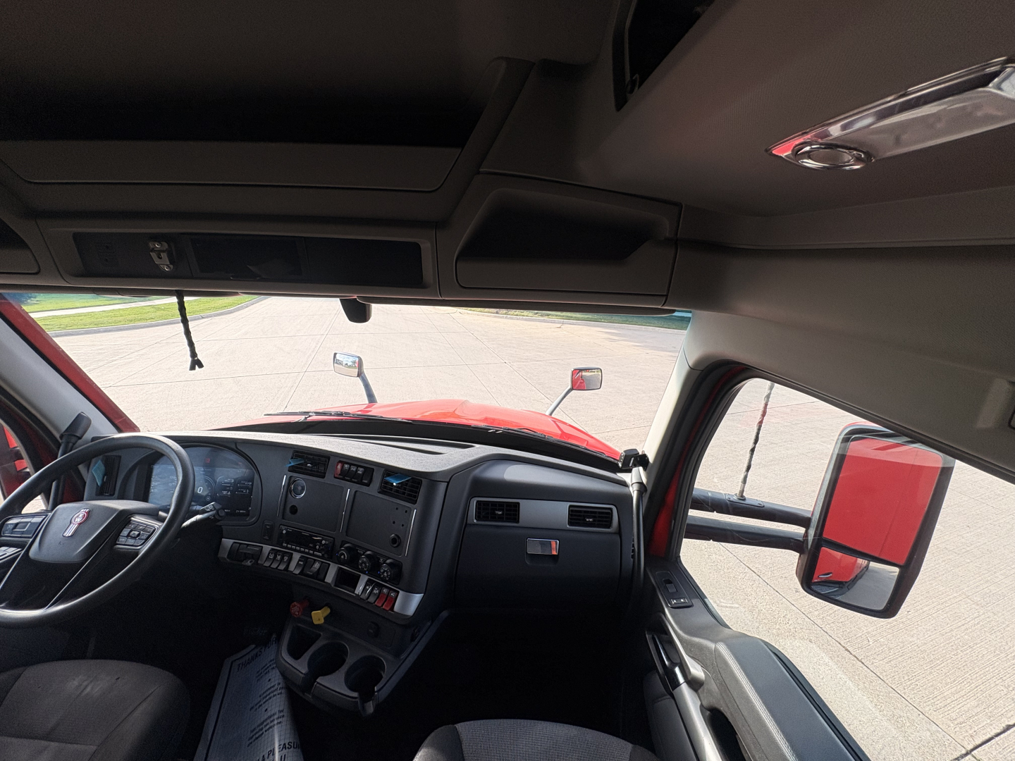 Interior of Kenworth T680 cab showing dashboard and steering wheel, with wide windshield and driver-side door mirror visible.