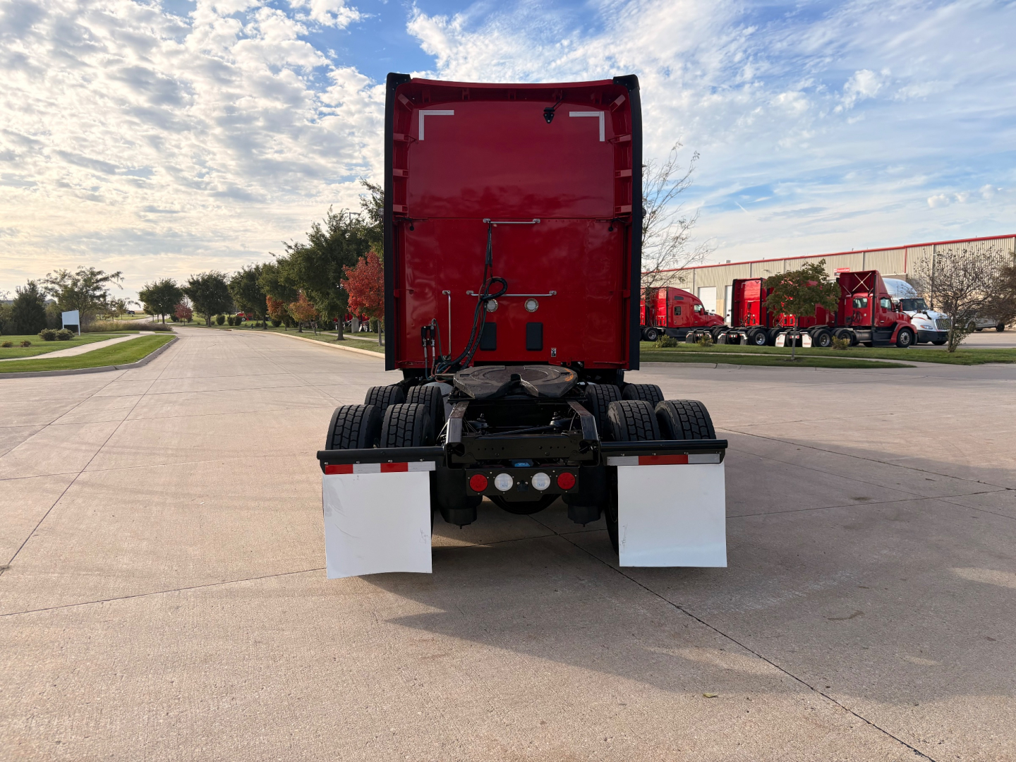 Rear view of a red Kenworth truck with gray mud flaps, exposed drivetrain, and black chassis in a truck lot.