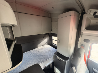 Kenworth T680 sleeper cab interior showing bunk bed with quilted mattress, overhead storage, and refrigerator unit.