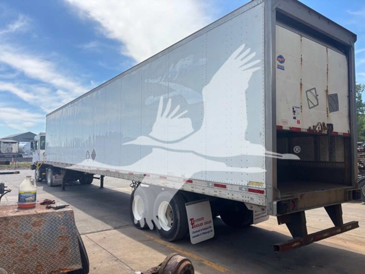 Trailer side view showing a white Utility 4000D-X box trailer with tandem axles and a roll-up rear door. VIN tag not visible.