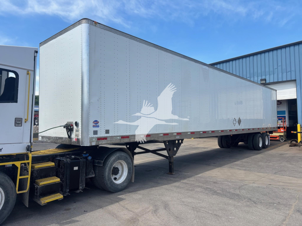 Trailer shows a long enclosed utility van trailer attached to a truck. Side features white graphic of flying birds and some hazard placards near the rear. Visible text includes no specific model text on the body, only general trailer markin