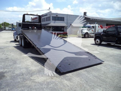 Truck scene showing a 2015 Peterbilt 348 rollback with a long lowered ramp in the foreground. Visible are other trucks in the lot and a white building in the background.