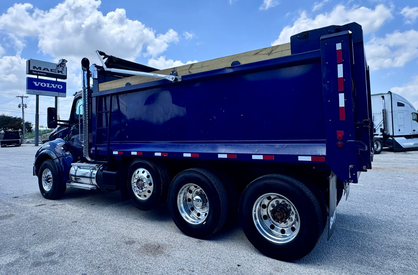 Kenworth T880 dump truck with blue dump bed, three rear wheels visible, chrome exhaust stack, and white cab in a dealership lot.