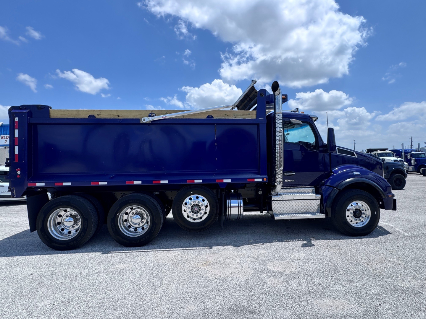 Kenworth T880 dump truck side view with dark blue cab and dump body, chrome fuel tanks and wheels, clear sky.