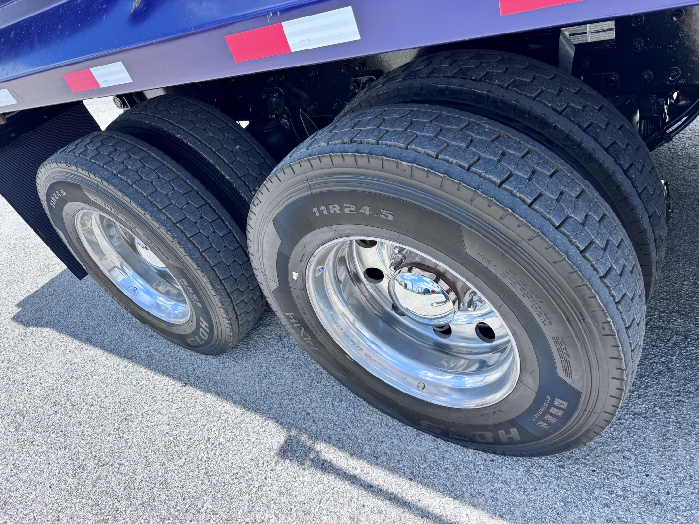 Truck wheel assembly with dual rear tires and chrome rims; tire sidewall shows markings 11R24.5 and other text; part of dump trailer visible.