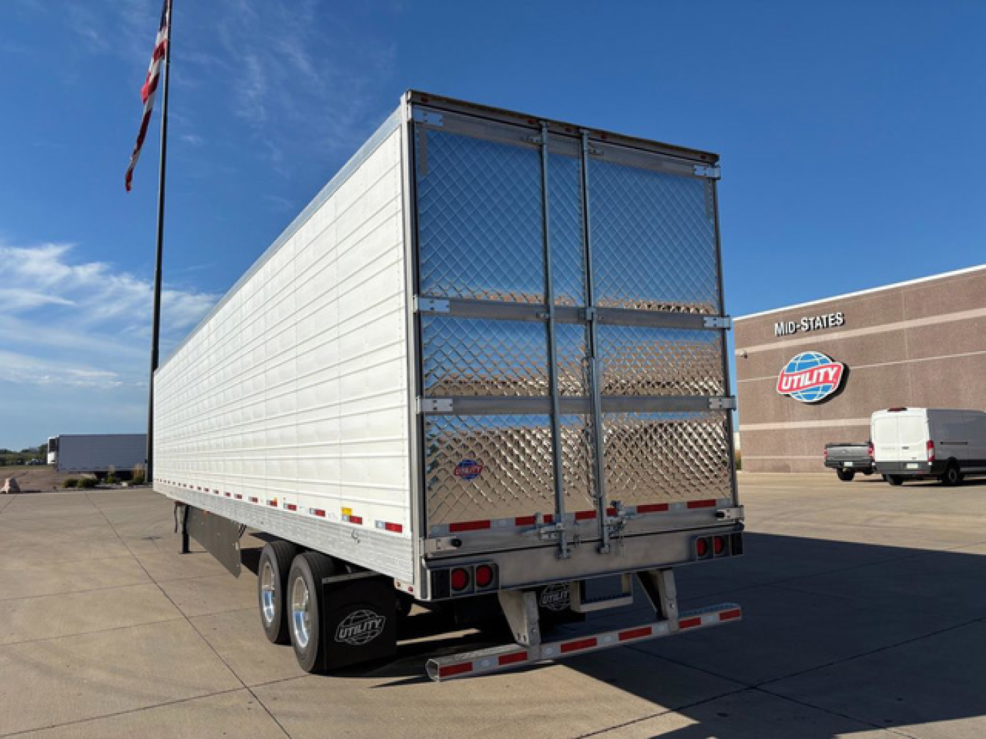 Rear view of a new 2027 Utility reefer trailer with diamond plate rear doors and dual rear axles, side walls white, U.S. flag pole in lot.