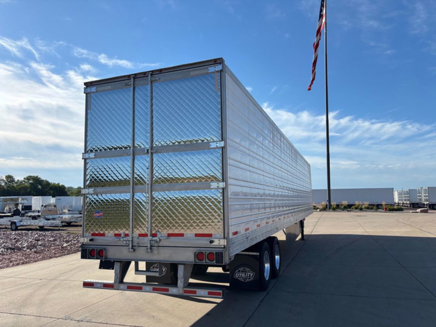 Utility refrigerated trailer rear view with diamond plate aluminum sides, dual rear doors, and red/white reflective tape. US flagpole and clear blue sky in background.
