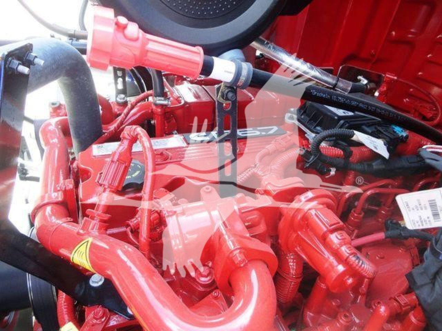 Engine bay of a 2027 Mack MD6 rollback truck showing red-painted components, hoses, and wiring with visible labels and a coolant reservoir cap.