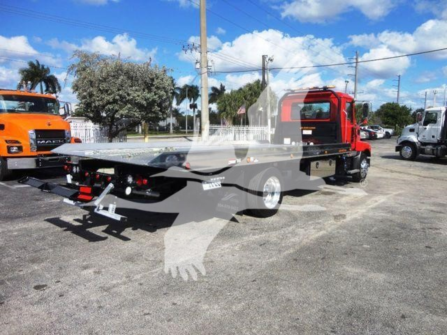 Tow truck chassis shown from rear-left side with a flatbed, red cab, black flatbed, and towing equipment; background includes other trucks and palm trees.