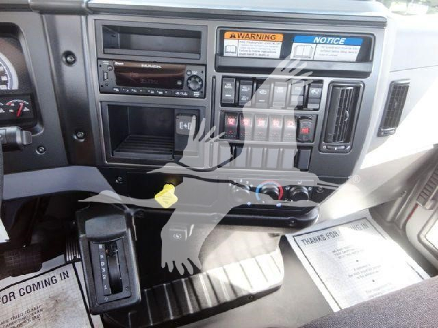 Cab interior showing dashboard with Mack logo and multiple controls, warning/notice stickers, radio, and climate controls.