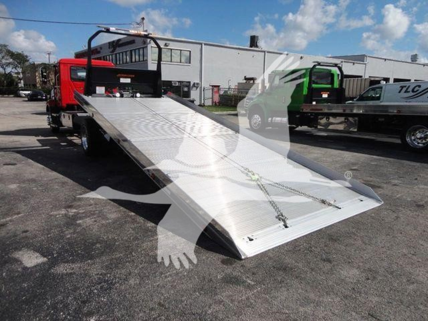 Flatbed tow truck bed with aluminum ramp and rear deck, shown in outdoor lot with other trucks; no visible text readable on components.