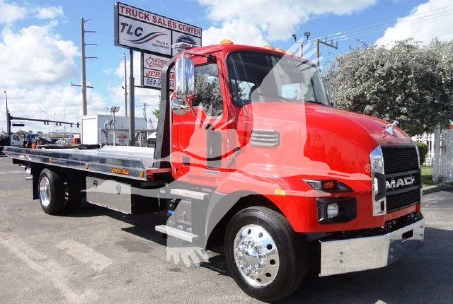 Flatbed tow truck in red Mack MD6 side view showing carrier bed and front grille with Mack logo; dealership lot in background.