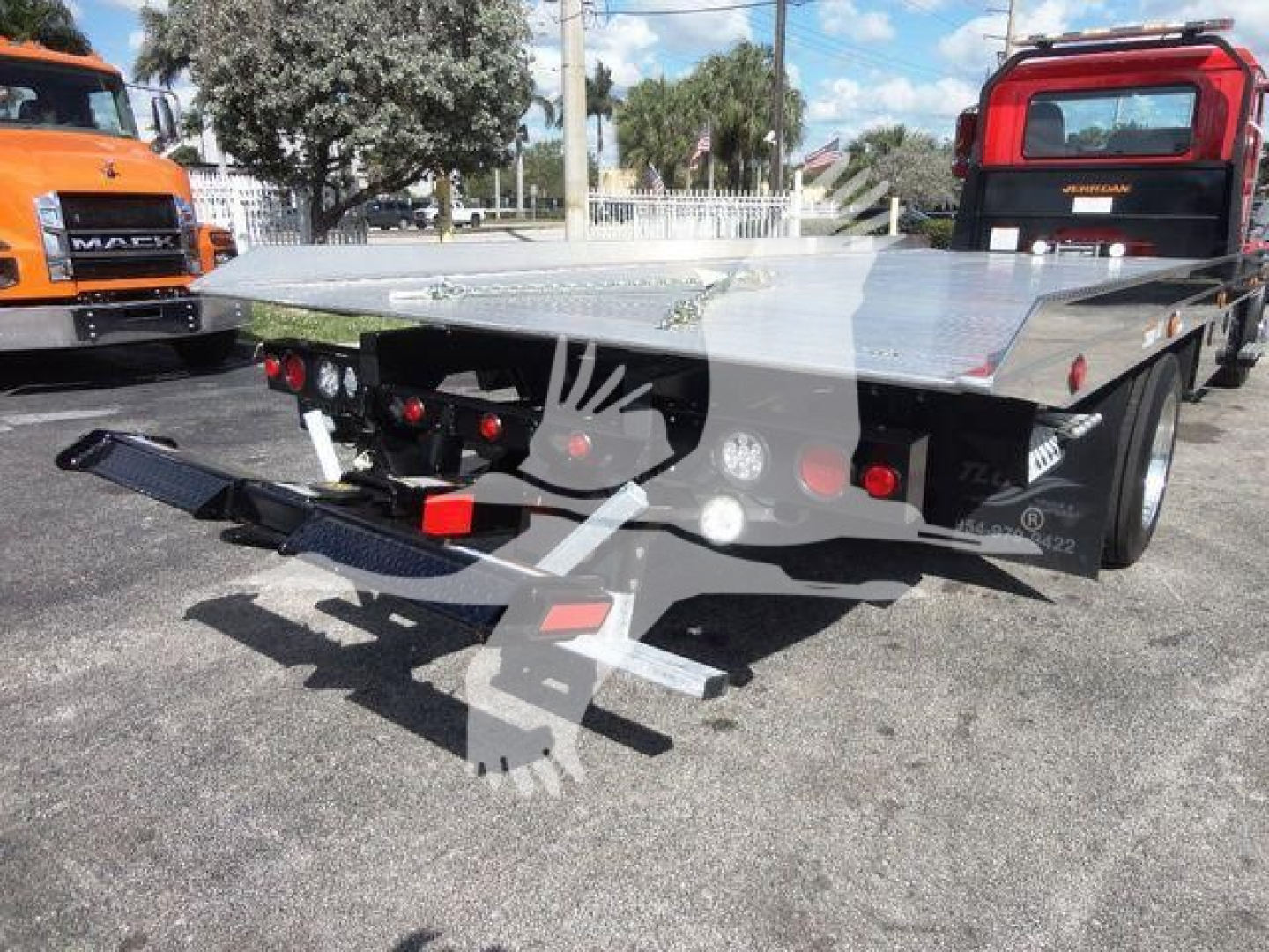 Flatbed tow truck rear view showing aluminum platform with ramps and taillights, Mack Md6? style truck in background.