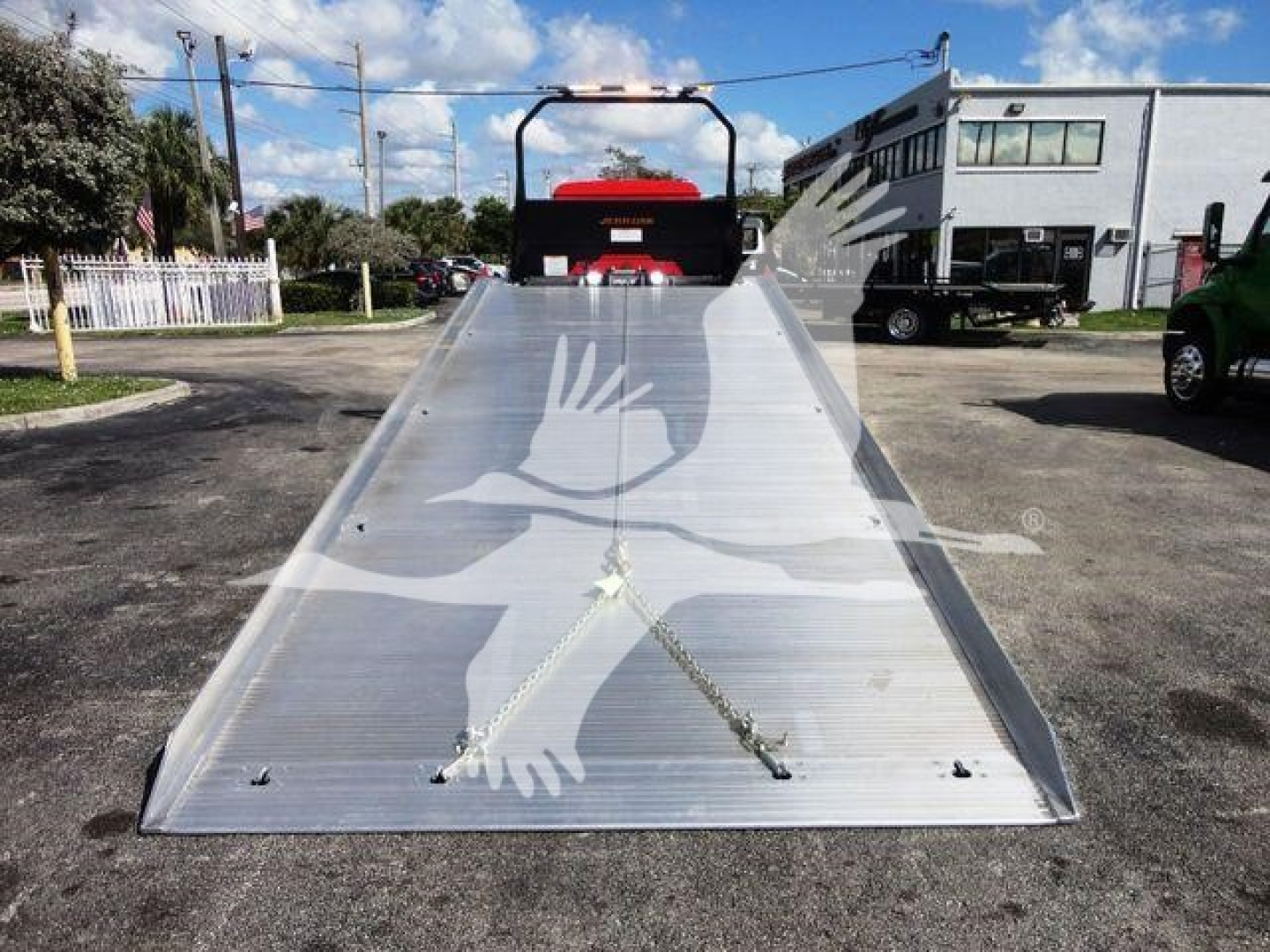 Truck ramp bed of a 2027 Mack MD6 rollback showing aluminum loading ramp with tie chains and a red cab in background.