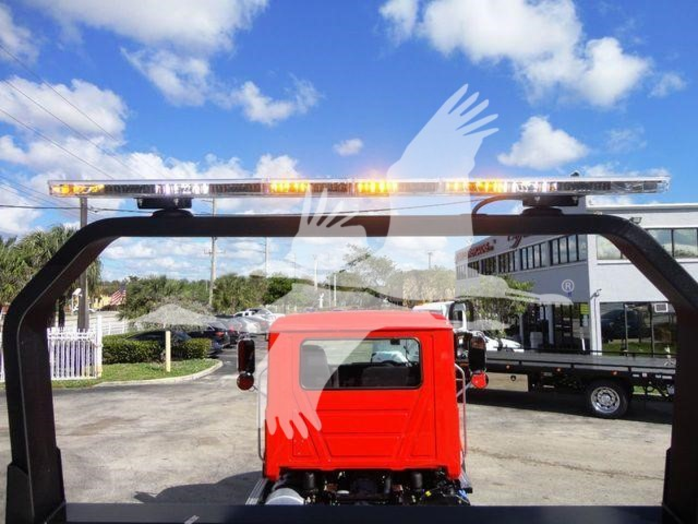 Flatbed view of a Mack MD6 rollback truck with amber warning light bar mounted on a frame, red cab, and blue sky background.