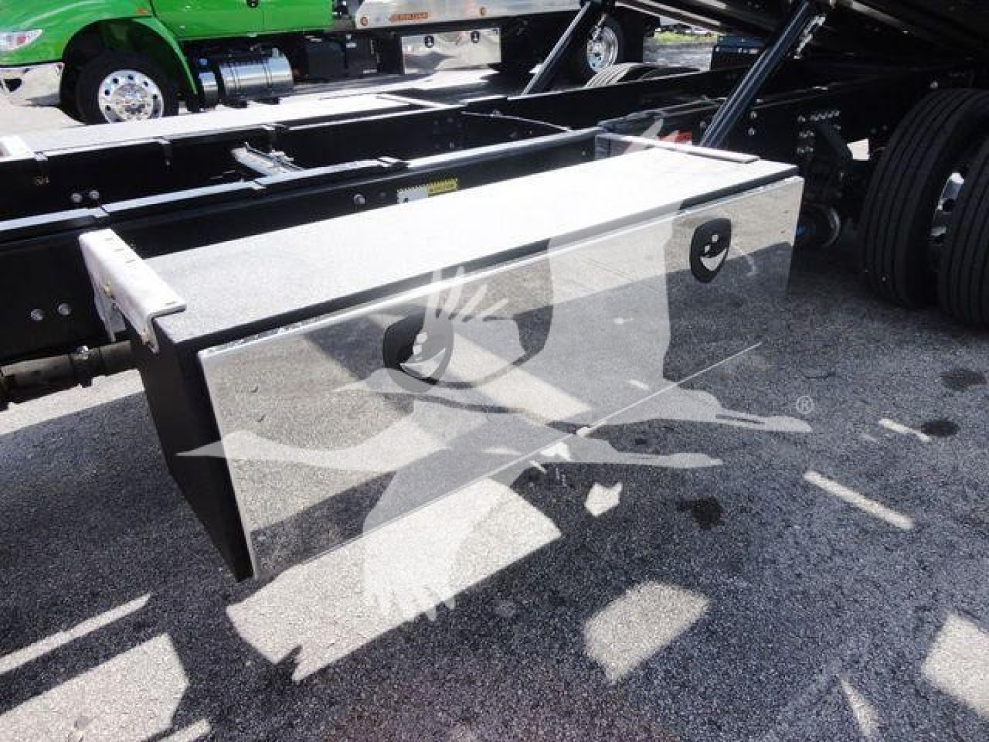 Flat metal toolbox/storage box mounted under a Mack MD6 truck frame, with supports and wheel well nearby.