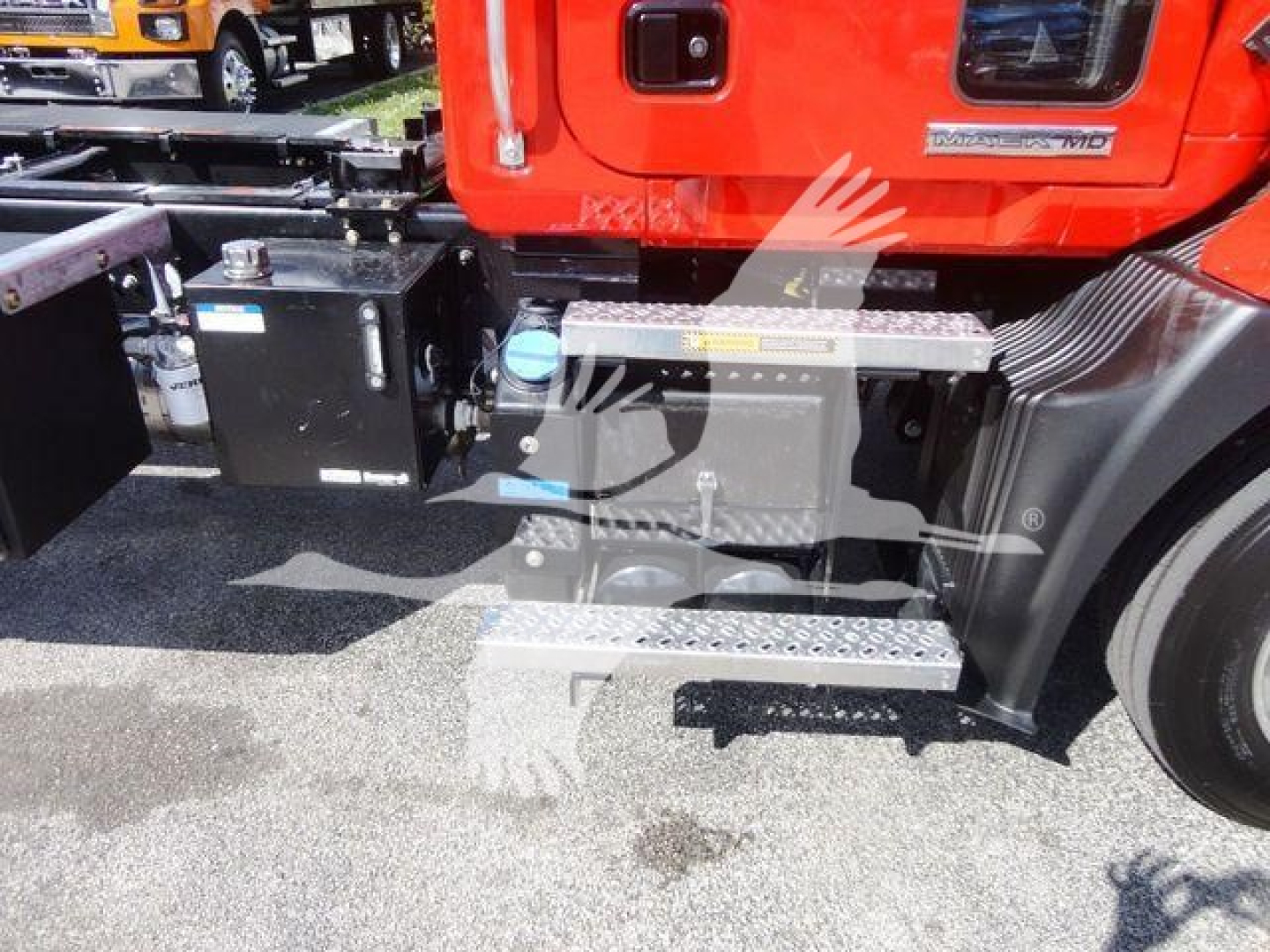 Truck side showing fuel tank and step with blue cap and warning labels, part of a red Mack MD series cab; visible stairs and battery box area.