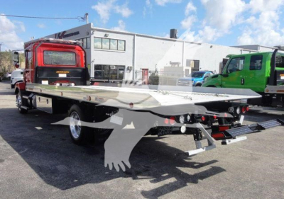 Flatbed rollback truck with long aluminum bed and rear-loading ramps visible; rear lights and dual wheels; dealership lot in background.