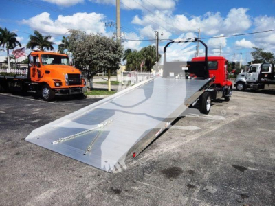 Flatbed rollback truck with a large aluminum ramp bed, tag gate visible, parked lot with other trucks.