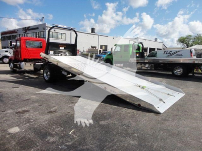 Delta-plateau flatbed tow truck bed attached to red tow truck in a dealership lot with other trucks in background.