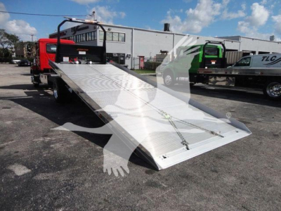 Flatbed tow truck bed with aluminum ramp and rear deck, shown in outdoor lot with other trucks; no visible text readable on components.