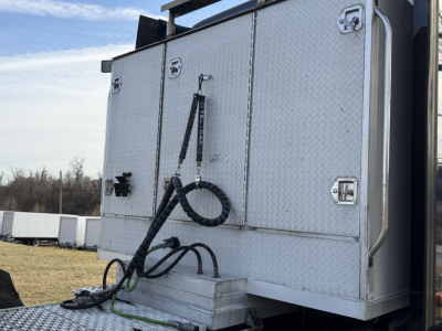 Truck rear with diamond plate storage box and hoses/coiled cable connected to auxiliary equipment; multiple latches on the storage compartments; metal exterior.