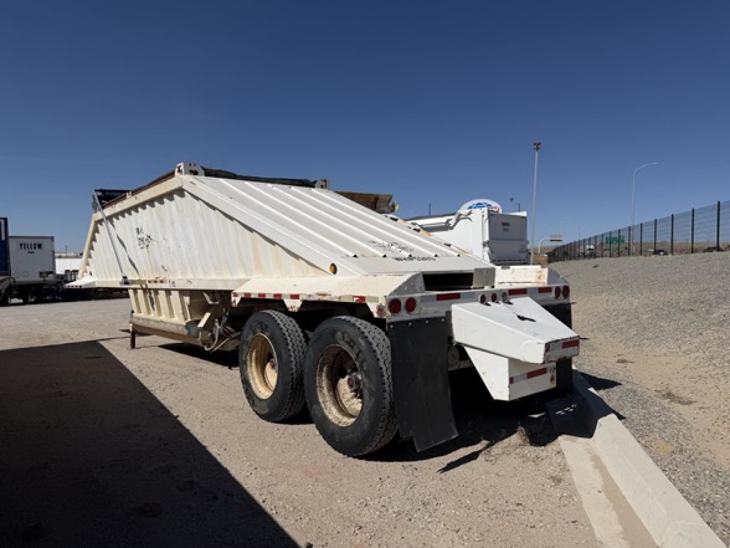 Construction trailer specialist air ride steel bottom dump trailer, rear view showing dual rear axles, drop-down tailgate, white paint, and gravel lot.