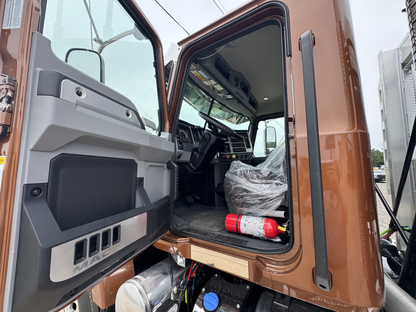 Truck cab interior visible through open door of a new Mack PI64T daycab with exposed dashboard, steering wheel, and plastic-covered seat; driver’s door open.