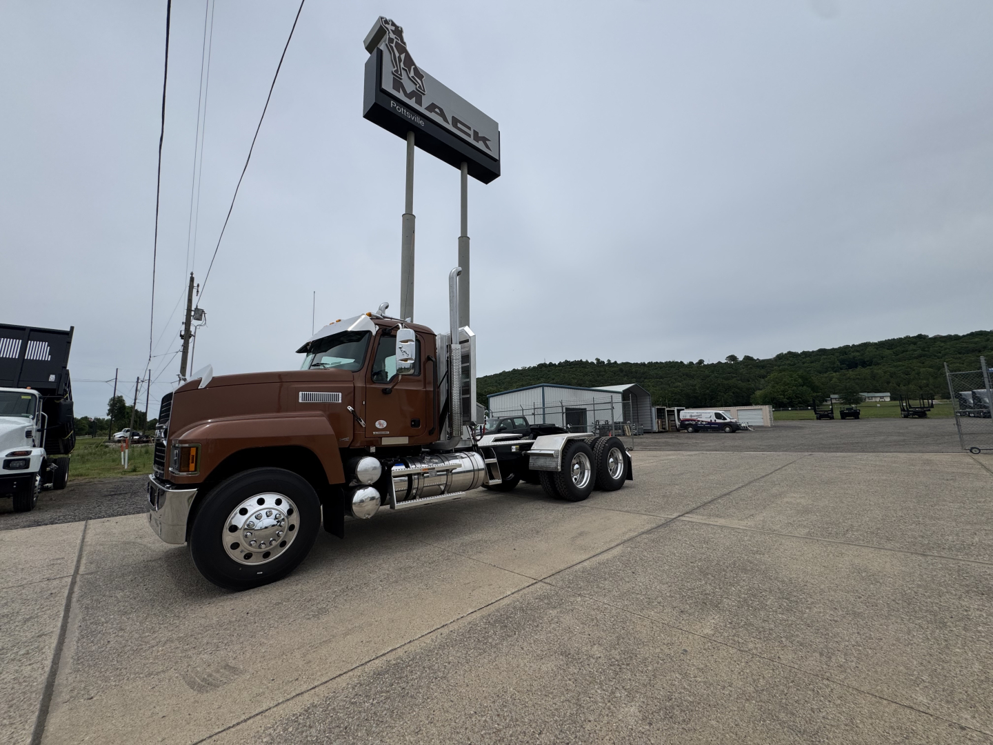 2027 Mack PI64T conventional daycab truck in brown with long hood, dual rear axles, parked in lot under dealership sign.
