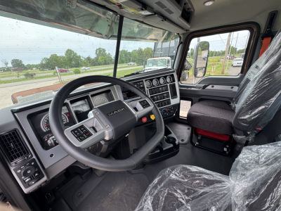 Dashboard and steering wheel of a 2027 Mack PI64T truck cab interior with plastic seat covers and control panel, showing steering wheel, instrument cluster, and overhead console.