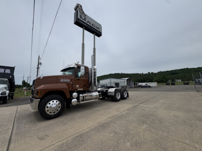 2027 Mack PI64T conventional daycab truck in brown with long hood, dual rear axles, parked in lot under dealership sign.