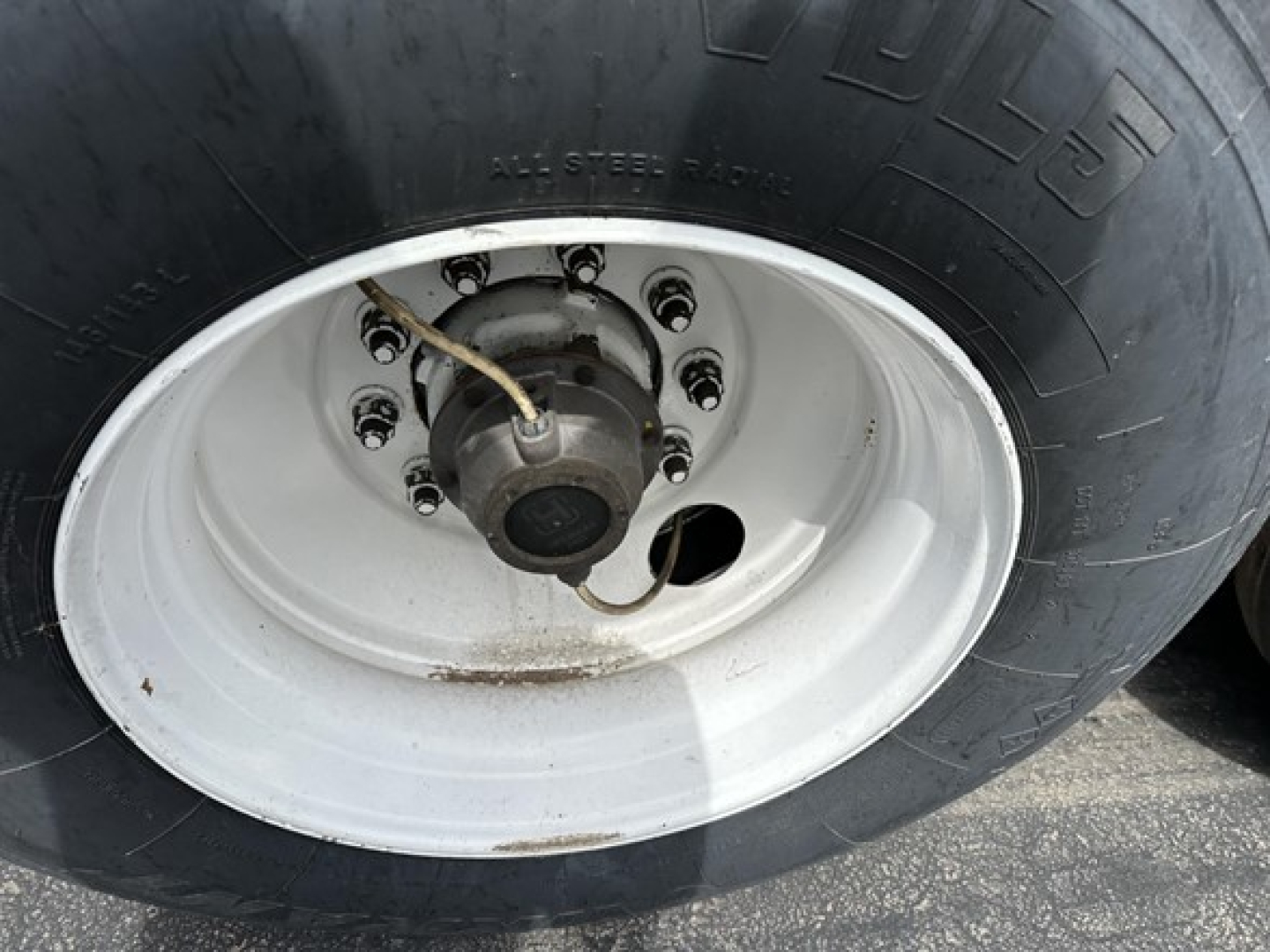 Trailer wheel showing large black tire with white steel rim and hub, multiple lug nuts, and a white paint hub area.