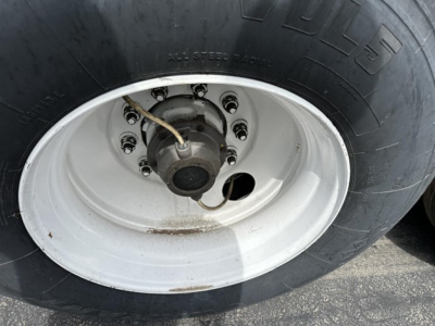 Trailer wheel showing large black tire with white steel rim and hub, multiple lug nuts, and a white paint hub area.