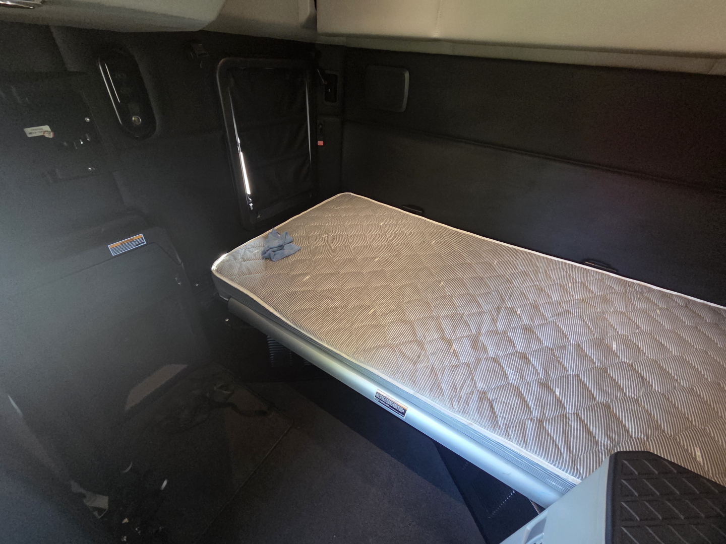 Kenworth T680 sleeper cab interior with a folded single-bed mattress on a platform, side controls and dark panels visible.
