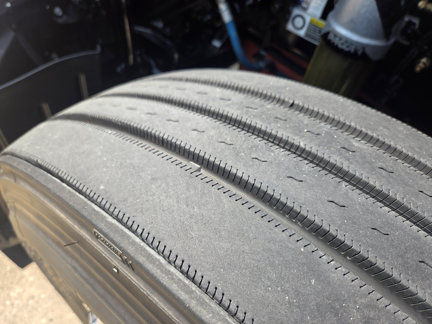 Tire tread close-up on Kenworth T680 truck wheel, showing parallel grooves and wear pattern; part of undercarriage visible in background.