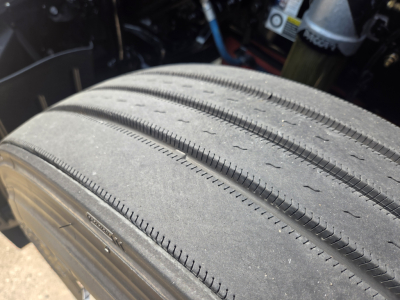 Tire tread close-up on Kenworth T680 truck wheel, showing parallel grooves and wear pattern; part of undercarriage visible in background.