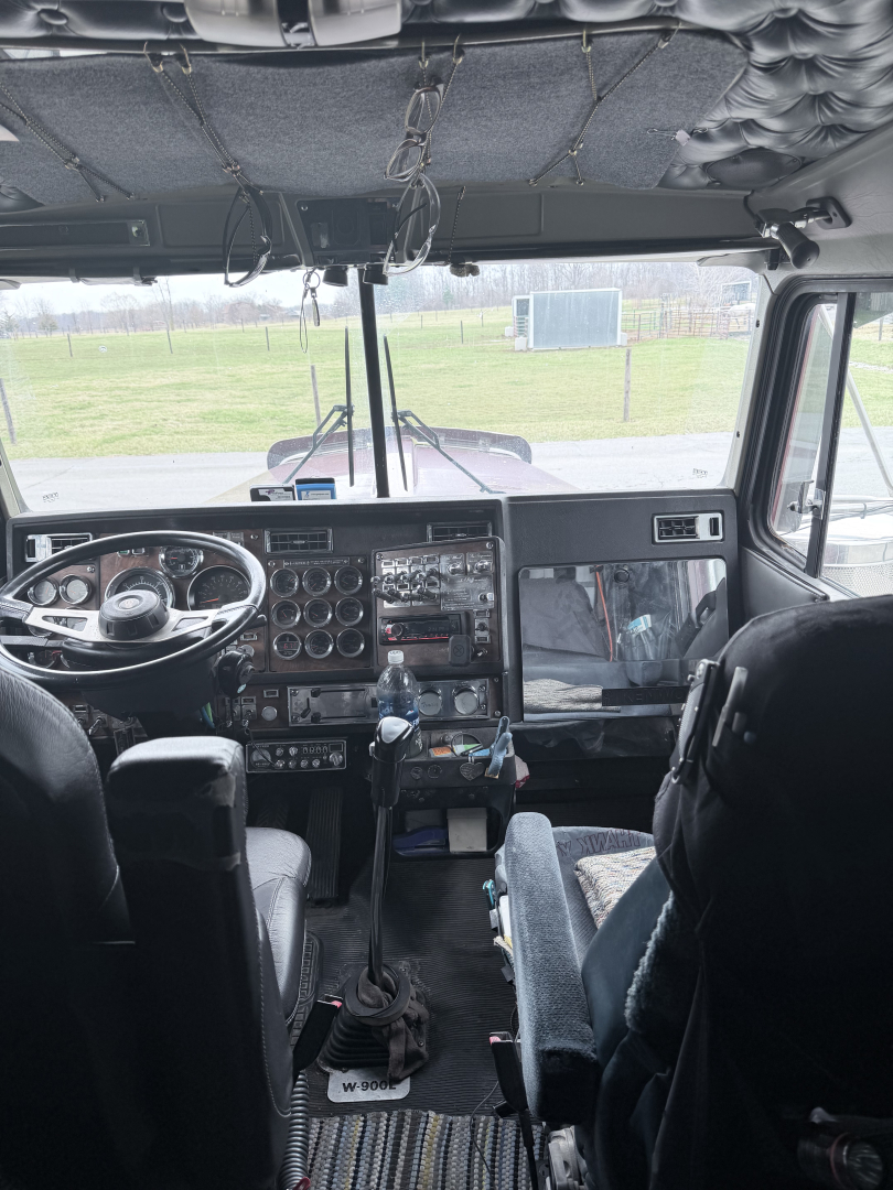 Kenworth W900L cab interior showing steering wheel, dashboard with gauges, gear shifter, and large windshield; overhead insulation and wiring visible.