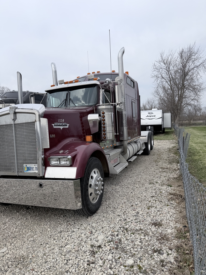 Kenworth W900L conventional sleeper truck in a used condition with chrome bumper, tall exhaust stacks, maroon cab, and trailers visible in background.