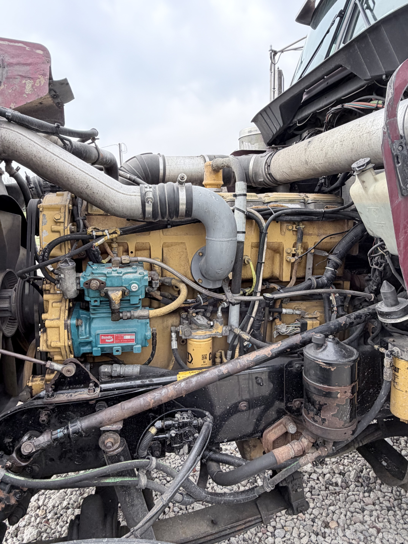 Kenworth W900L engine bay showing yellow engine block, blue fuel pump, hoses, and surrounding mechanical components.