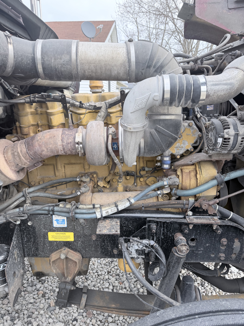 Truck engine bay of a 1999 Kenworth W900L showing large turbo intake hose, yellow engine block, various hoses and wiring, and metal components.