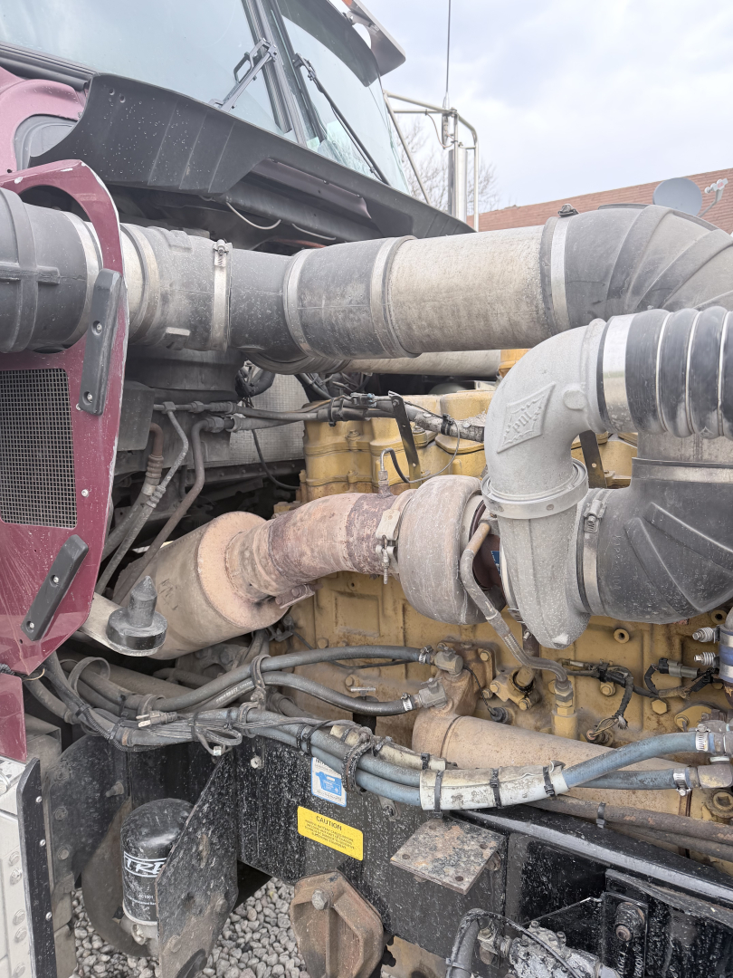 Engine bay view of a 1999 Kenworth W900L showing diesel engine and large intake hoses, visible turbocharger, coolant hoses, and surrounding components.