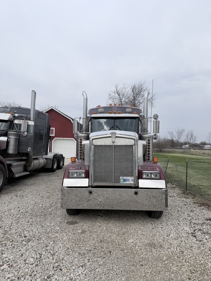 Front view of a 1999 Kenworth W900L conventional sleeper truck showing a large chrome grille, dual tall exhaust stacks, amber clearance lights, and a front bumper. License plate visible on bumper.