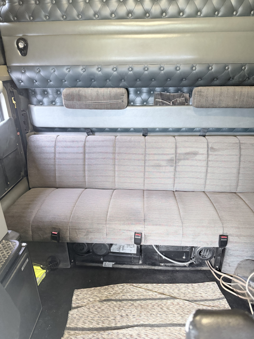 Kenworth W900L interior sleeper cab side view showing grey upholstered bench seat, quilted padded wall, storage pockets, and metal framework.
